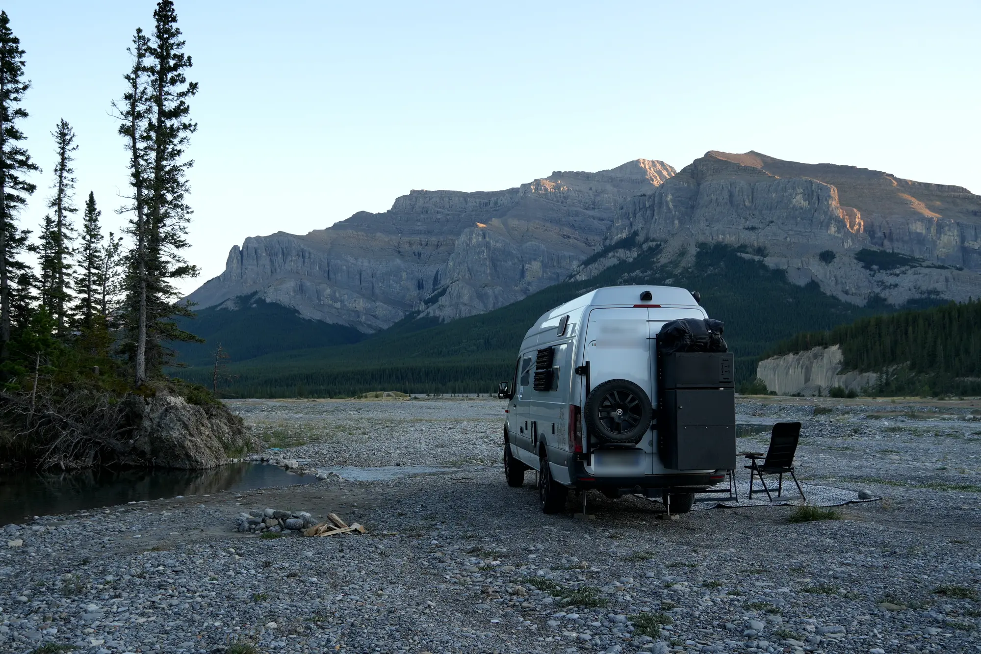 Finn parked alone on a gravel riverbed below a forested mountain, a camp chair beside it